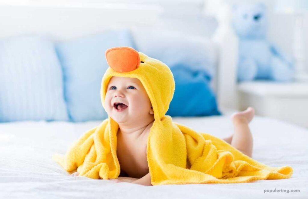A Baby In A Yellow Duck Towel Laying On A Bed.	