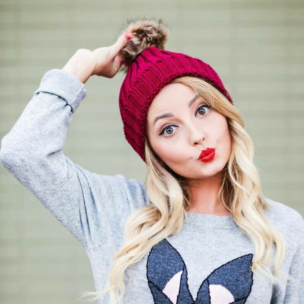 a blonde woman wearing a burgundy sweater and pom pom beanie.	